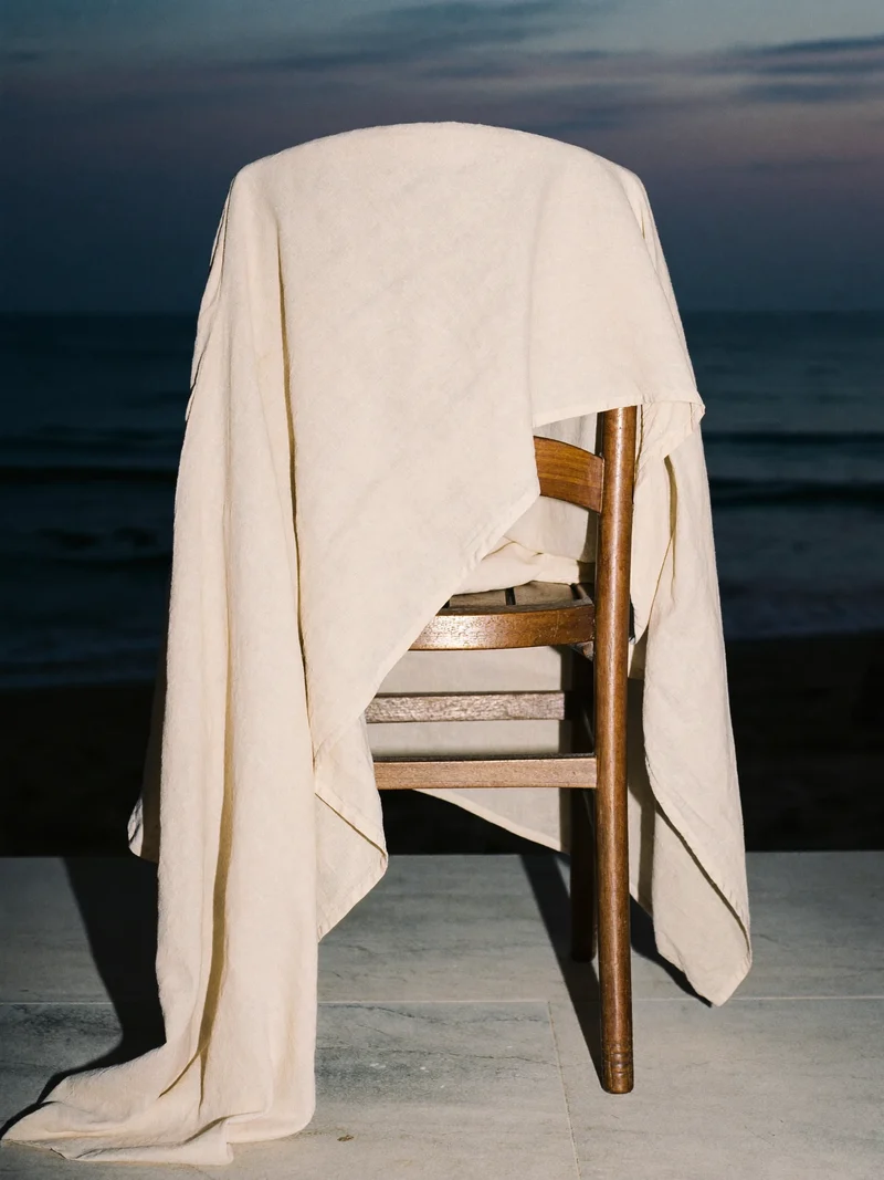 Sandfarbener Leinenkaftan über einem Holzstuhl auf einer Terrasse am Abendmeer