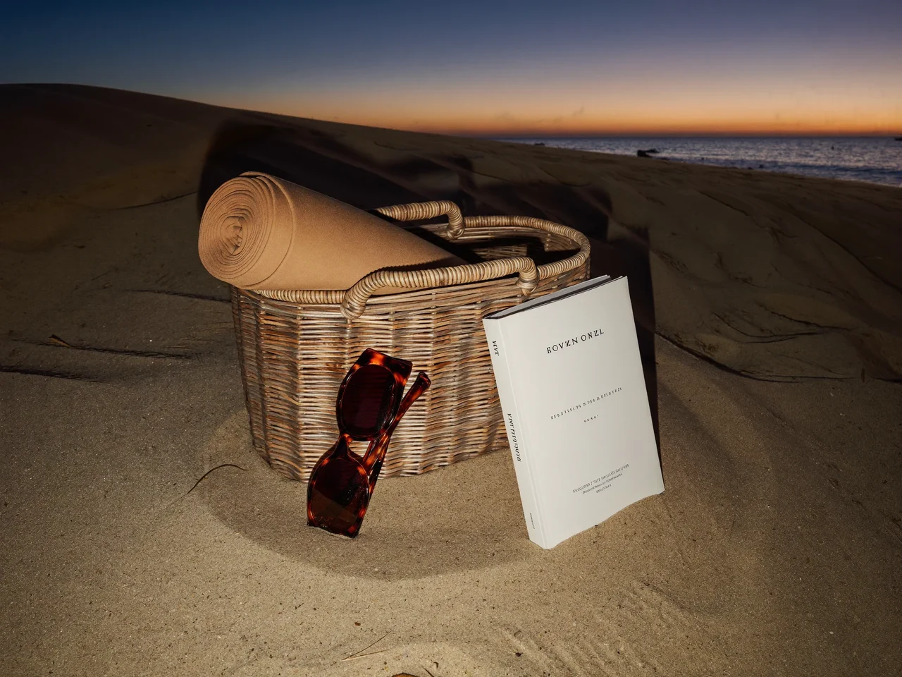 Korbtasche mit Sarong, Sonnenbrille und Taschenbuch auf Sand in der Dämmerung